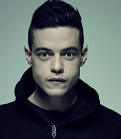 A man with a serious expression, short dark hair, and wearing a black hoodie. This is Elliot Alderson from the Netflix show Mr. Robot, known for his intense gaze and sharp features, creating a striking and memorable impression.