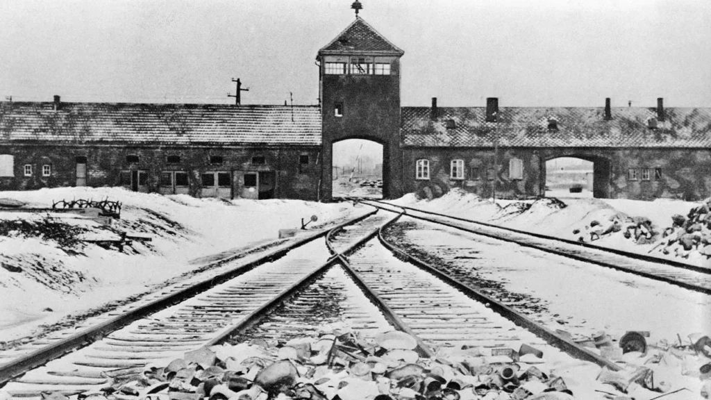 Black-and-white photograph of the main entrance to Auschwitz-Birkenau, a Nazi concentration and extermination camp. The image shows snow-covered train tracks leading directly into the gatehouse, a central brick structure with an archway and a watchtower above. On either side of the gatehouse are single-story brick buildings, and the foreground is littered with discarded belongings, symbolizing the atrocities committed at the camp.