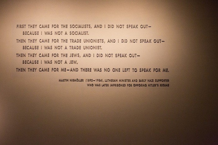 The image shows a wall with Martin Niemöller's famous quote, written in bold, clear lettering. The text reads: "First they came for the Socialists, and I did not speak out—because I was not a Socialist. Then they came for the Trade Unionists, and I did not speak out—because I was not a Trade Unionist. Then they came for the Jews, and I did not speak out—because I was not a Jew. Then they came for me—and there was no one left to speak for me." Below the quote, there is an attribution: Martin Niemöller (1892–1984), Lutheran Minister and early Nazi supporter who was later imprisoned for opposing Hitler’s regime. The simple, solemn presentation emphasizes the profound and haunting message of the text, urging viewers to reflect on the consequences of silence and inaction in the face of injustice.